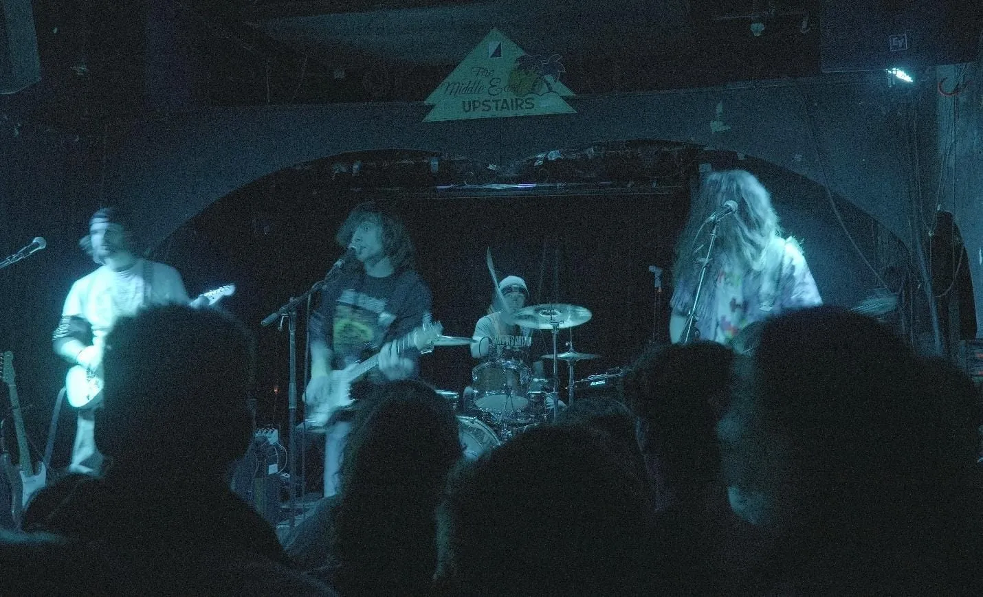 The image depicts a dimly lit indoor music venue with a band performing on stage, surrounded by a small audience.

AI-generated content may be incorrect.
